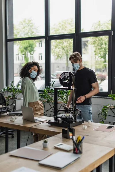 Interracial colleagues in medical masks standing near 3D printer in modern office — Stock Photo