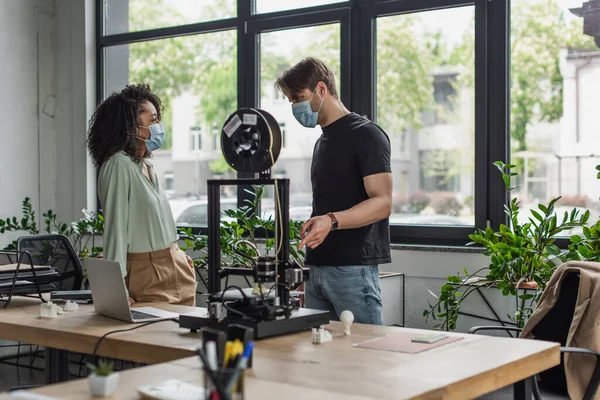 Diseñadores interraciales en máscaras médicas hablando cerca de la impresora 3D en la oficina moderna - foto de stock