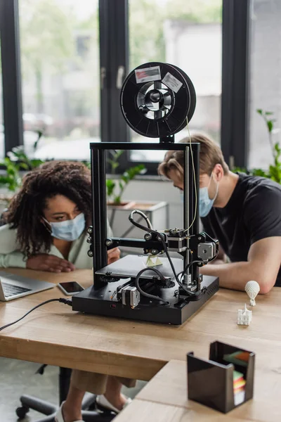 Interracial colleagues in medical masks working with 3D printer in modern office — Stock Photo