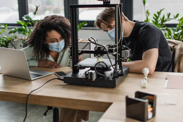 Interracial colleagues in medical masks looking at 3D printed model production in modern office — Stock Photo