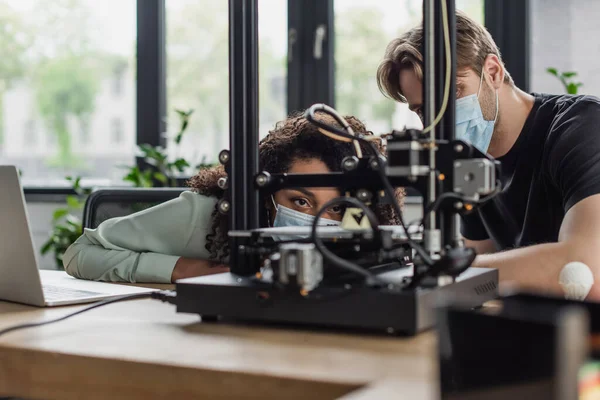 Designers interracial dans les masques médicaux regardant la production de modèles imprimés en 3D dans le bureau moderne — Photo de stock