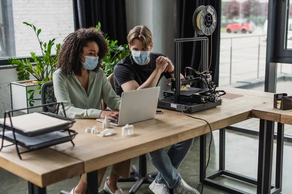 Interracial colleagues in medical masks working with laptop and 3D printer in modern office — Stock Photo