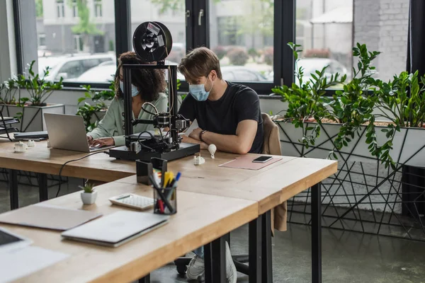 Interracial colleagues in medical masks typing using laptop near 3D printer in modern office — Stock Photo