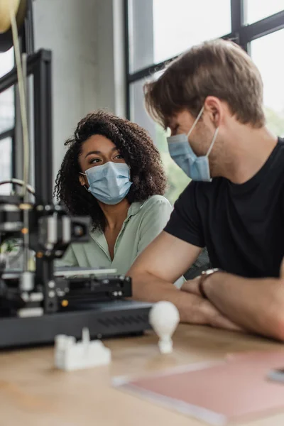 Interracial colleagues in medical masks sitting near 3D printer and plastic figures in modern office — Stock Photo