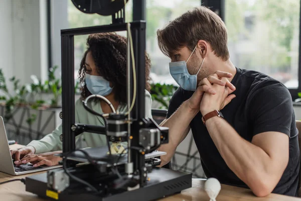 Межрасовые коллеги в медицинских масках, работающие с 3D принтером и ноутбуком в современном офисе — стоковое фото