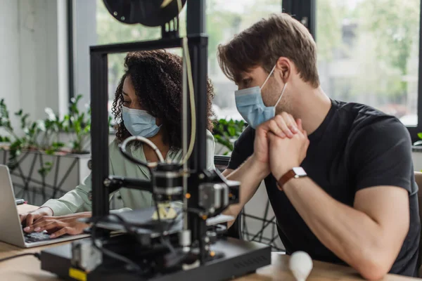 Collègues interracial dans des masques de protection travaillant avec un ordinateur portable près de l'imprimante 3D dans le bureau moderne — Photo de stock