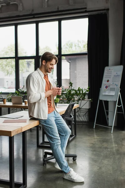 Giovane uomo sorridente in abiti casual che scrive sul cellulare in un moderno open space — Foto stock