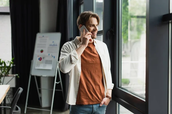 Улыбающийся молодой человек, говорящий по мобильному телефону возле флип-чарта с графиками и графиками в современном офисе — стоковое фото