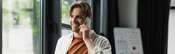 Giovane uomo sorridente in camicia beige che parla sul cellulare vicino a lavagna a fogli mobili con grafici in ufficio moderno, banner — Foto stock