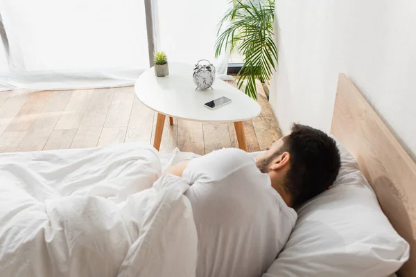 Vista posteriore di smartphone e sveglia sul tavolo vicino all'uomo che dorme sul letto — Foto stock