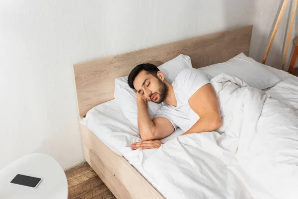 Hombre barbudo durmiendo en la cama cerca de teléfono inteligente con pantalla en blanco en la mesita de noche - foto de stock