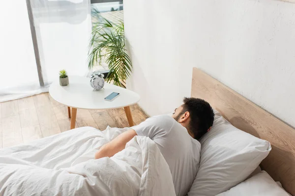 Vue arrière de l'homme dormant près du téléphone cellulaire et du réveil dans la chambre — Photo de stock