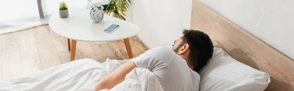 Вид сзади мужчины, спящего на кровати рядом с мобильным телефоном на тумбочке, баннер — стоковое фото