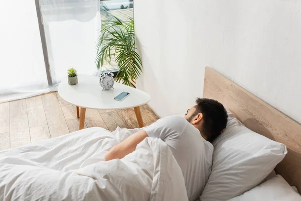 Rückansicht von Smartphone und Wecker auf dem Tisch neben schlafendem Mann — Stockfoto