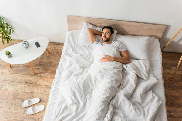Vista superior del hombre barbudo durmiendo en ropa de cama blanca cerca de teléfono inteligente y despertador - foto de stock