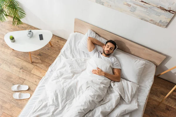 Vue du dessus de l'homme dormant sur le lit près des pantoufles et smartphone dans la chambre — Photo de stock