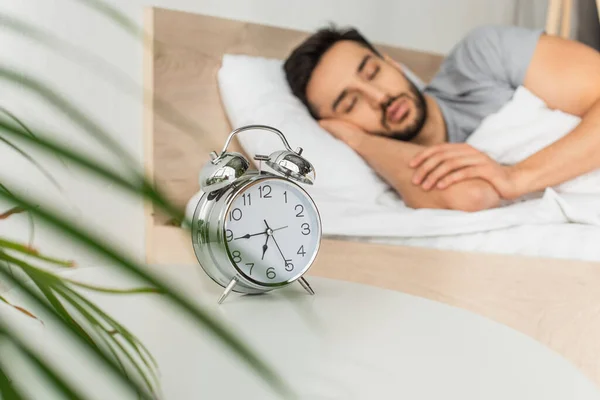 Reloj despertador cerca de la planta borrosa y el hombre durmiendo en la cama - foto de stock