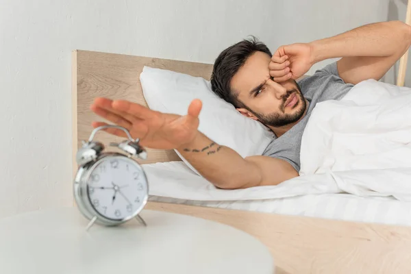 Young man pulling hand to blurred alarm clock while waking up — Stock Photo