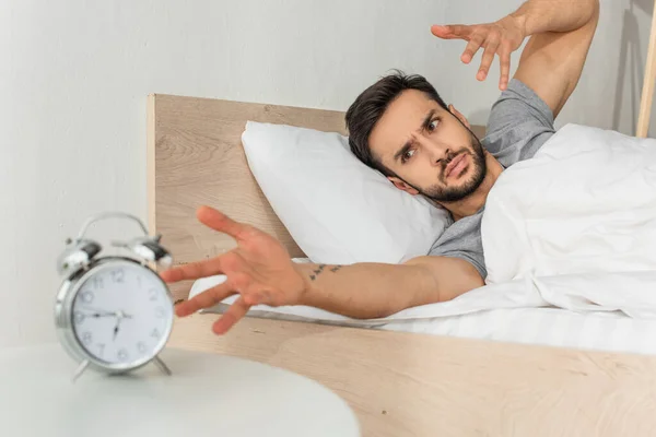 Homem irritado olhando para despertador desfocado na cama — Fotografia de Stock