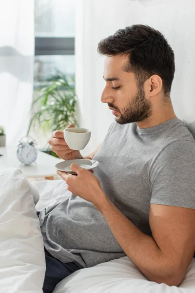 Side view of young man holding cup and saucer on bed — Stock Photo