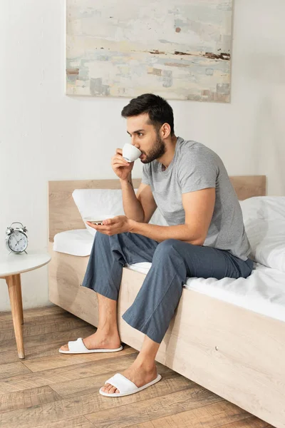 Бородатый мужчина в пижаме пьет кофе на кровати дома — Stock Photo