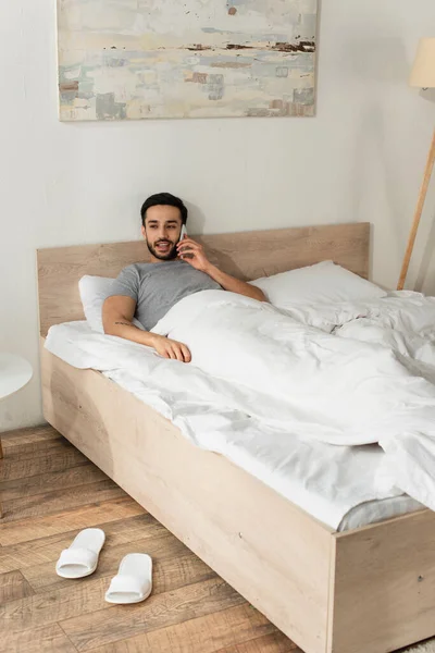 Uomo sorridente che parla su smartphone sul letto — Foto stock