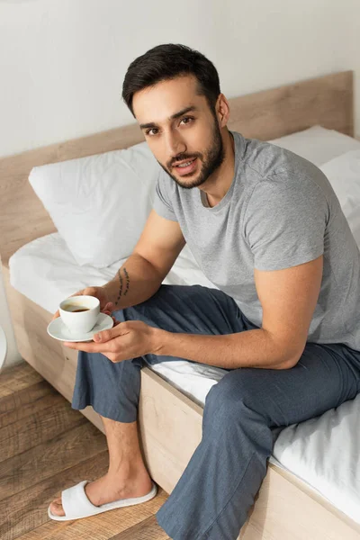 Молодой человек в пижаме держит блюдце и кофейную чашку на кровати — стоковое фото
