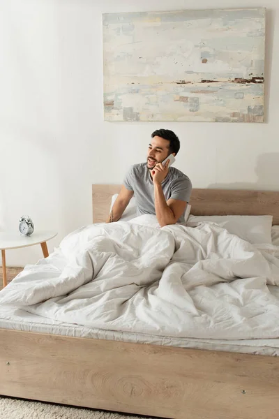 Uomo felice che parla sul telefono cellulare sul letto vicino sveglia in camera da letto — Foto stock