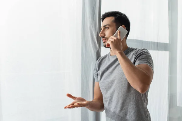 Vista lateral del hombre apuntando con la mano mientras habla en el teléfono inteligente cerca de la ventana en casa - foto de stock