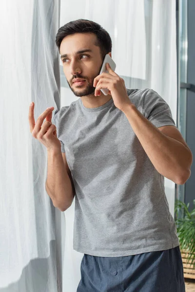 Young man in pajama talking on cellphone near window at home — Stock Photo