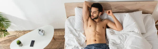 Top view of shirtless man lying near alarm clock and smartphone on bedside table, banner — Stock Photo