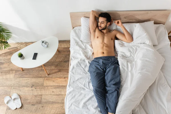 Vista superior del hombre sin camisa acostado en la cama cerca del teléfono inteligente con pantalla en blanco y reloj despertador en la mesita de noche - foto de stock