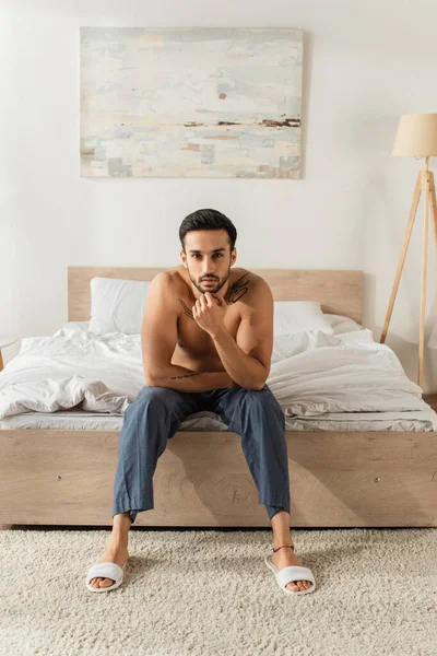 Чоловік без сорочки в тапочках і штанях дивиться на камеру на ліжку — стокове фото