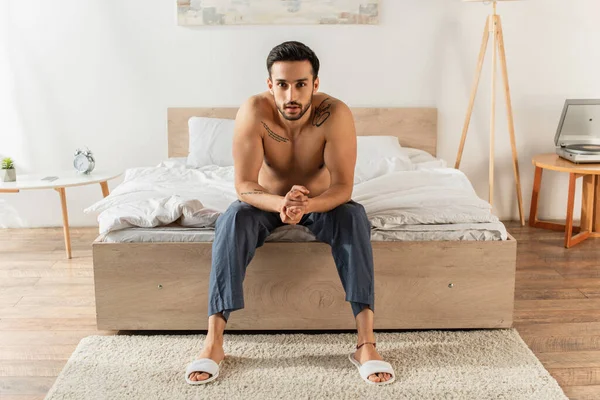 Jovem sem camisa homem sentado na cama e olhando para a câmera — Fotografia de Stock