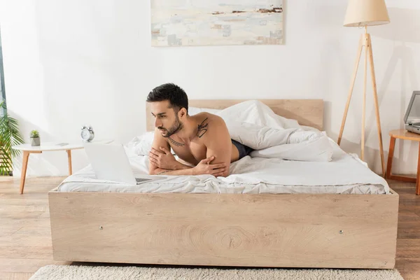 El hombre sin camisa mirando el ordenador portátil en la cama por la mañana - foto de stock
