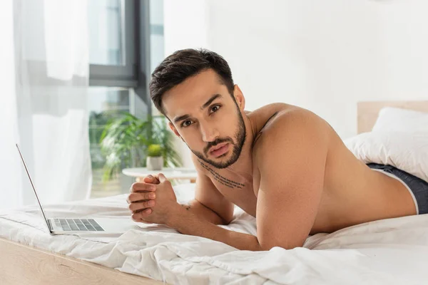 Мужчина без рубашки смотрит в камеру возле ноутбука на кровати дома — стоковое фото