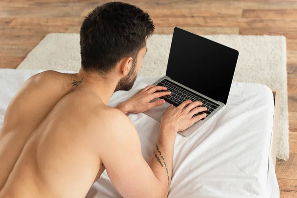 Vista aérea del hombre sin camisa usando el ordenador portátil con la pantalla en blanco en la cama - foto de stock