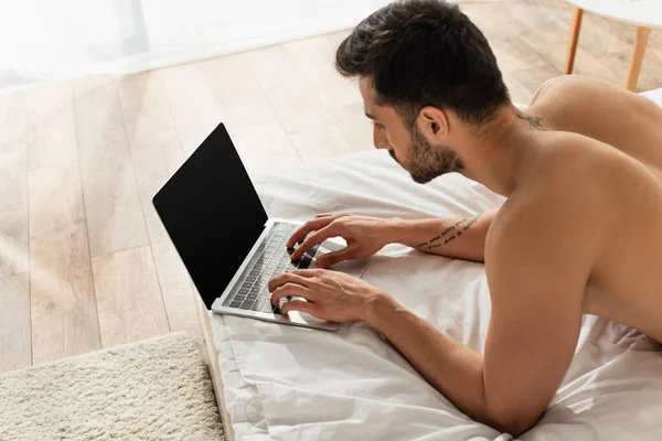 Vista de ángulo alto del hombre tatuado usando el ordenador portátil con la pantalla en blanco en la cama - foto de stock