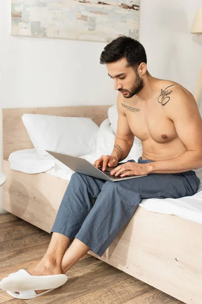 Uomo senza maglietta in pantofole e pantaloni utilizzando il computer portatile sul letto — Foto stock