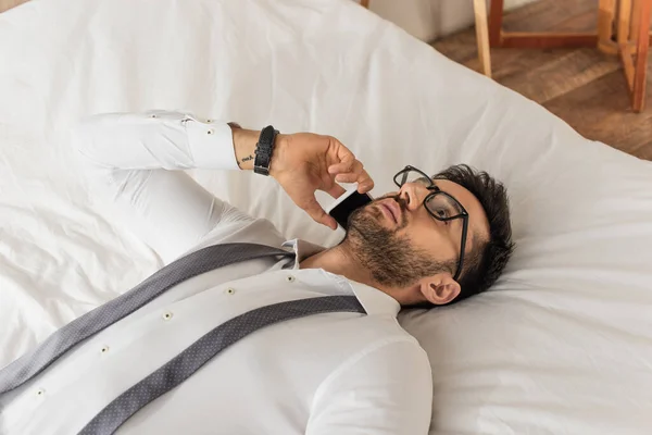 Homme d'affaires en lunettes et chemise parlant sur smartphone tout en étant couché sur le lit — Photo de stock