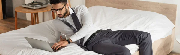 Empresário em óculos usando telefone celular e laptop na cama, banner — Fotografia de Stock