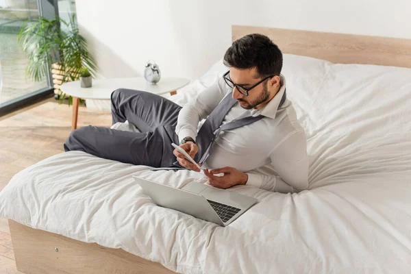 Businessman using smartphone near laptop on bed at home — Stock Photo