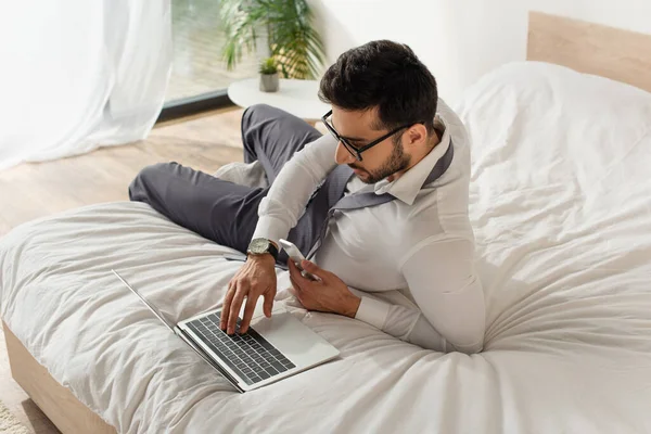 Young businessman in eyeglasses using laptop and holding smartphone on bed — Stock Photo