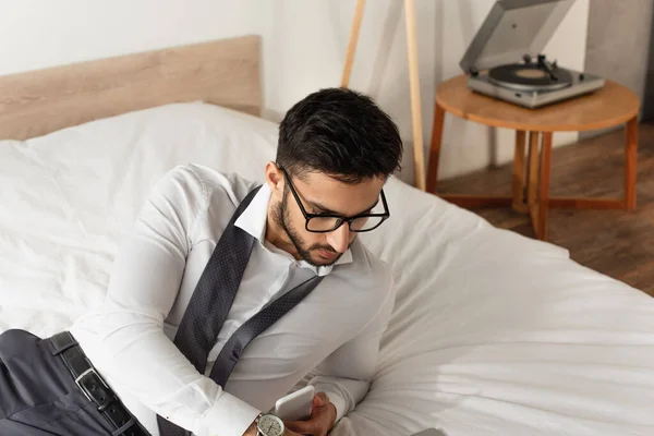 Uomo d'affari con gli occhiali che tiene il cellulare sul letto — Foto stock