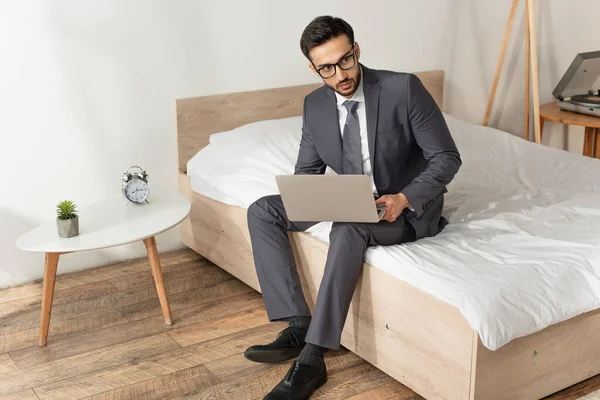 Businessman in eyeglasses holding laptop on bed near alarm clock on bedside table — Stock Photo