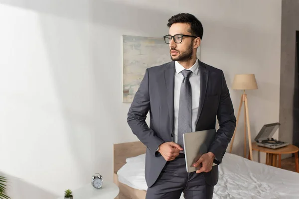 Junger Geschäftsmann in Anzug und Brille hält Laptop im Schlafzimmer — Stockfoto