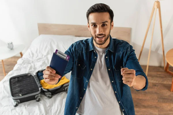 Emocionado hombre con pasaporte y mostrando sí gesto cerca de la maleta borrosa en la cama - foto de stock