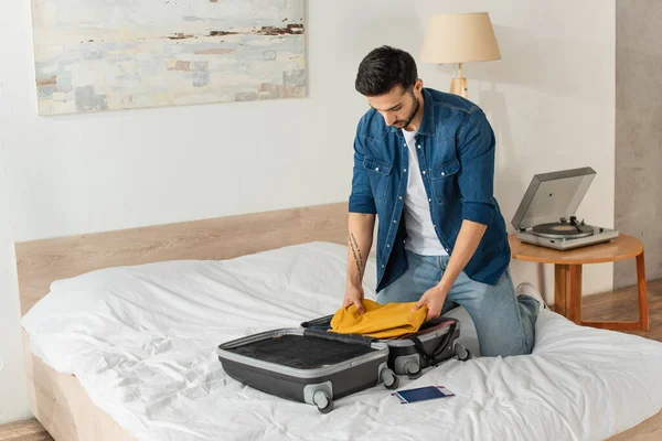 Bearded man putting clothes in suitcase near passport on bed — Stock Photo