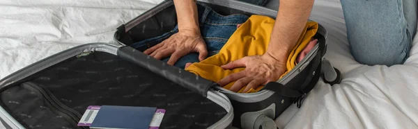 Vista recortada del hombre que presiona la ropa en la maleta cerca del pasaporte y billete de avión en la cama, pancarta - foto de stock