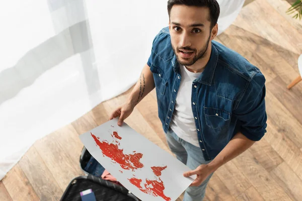 High angle view of smiling traveler holding map near blurred suitcase at home — Stock Photo